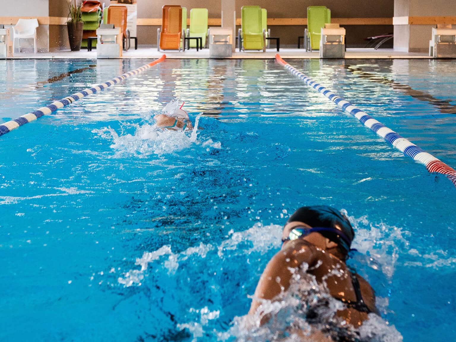 Zwei Schwimmerinnen ziehen ihre Bahnen im großen Innenbecken, am Kopfende stehen Liegestühle..