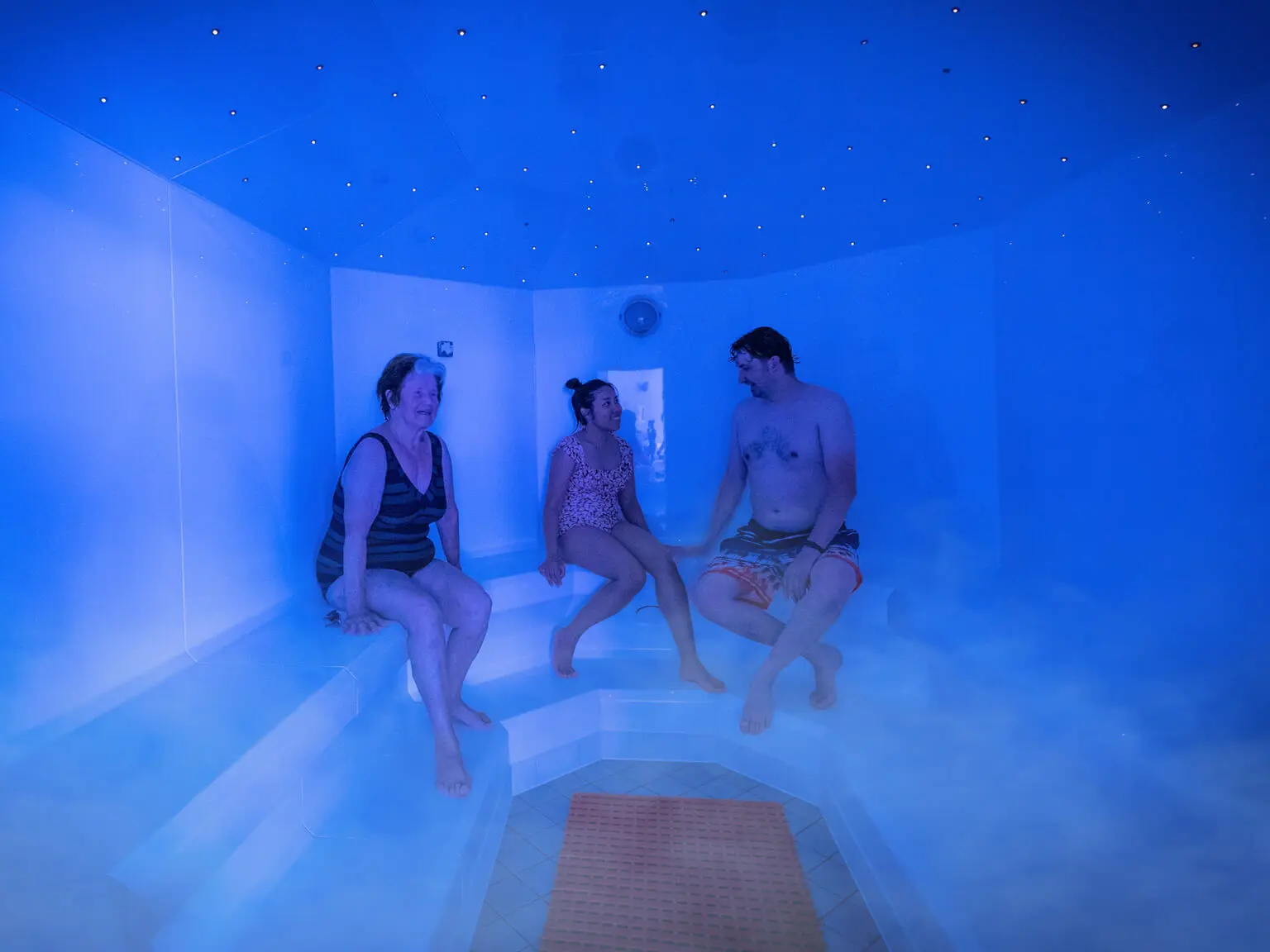 Drei Personen sitzen im Dampfbad mit kleinen leuchtenden Lichtern an der Decke.