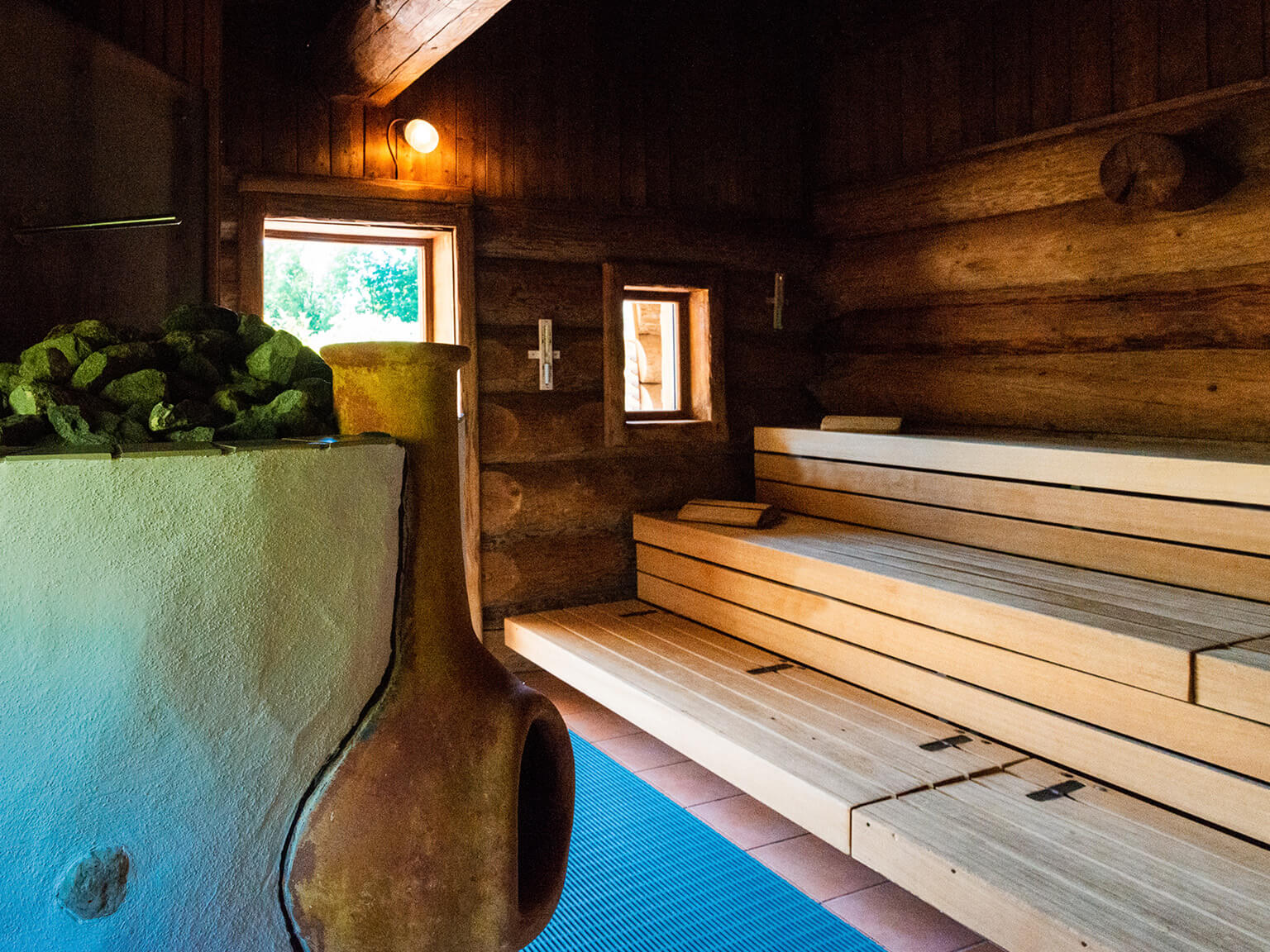 Blick in eine Sauna. Rechts die Sitzbänke aus hellem Holz, links der Ofen mit kleinem Kamin