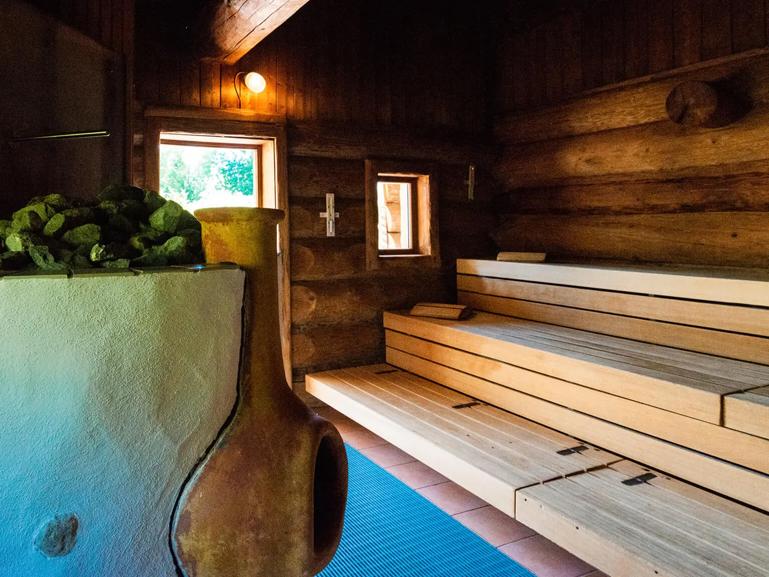 Blick in eine Sauna. Rechts die Sitzbänke aus hellem Holz, links der Ofen mit kleinem Kamin