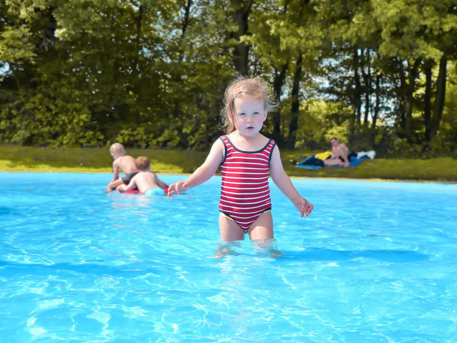 Ein kleines Mädchen steht im Wasser des Planschbeckens. Links dahinter spielen zwei Jungs im Wasser. Auf der Wiese im Hintergrund liegt ein Erwachsener.