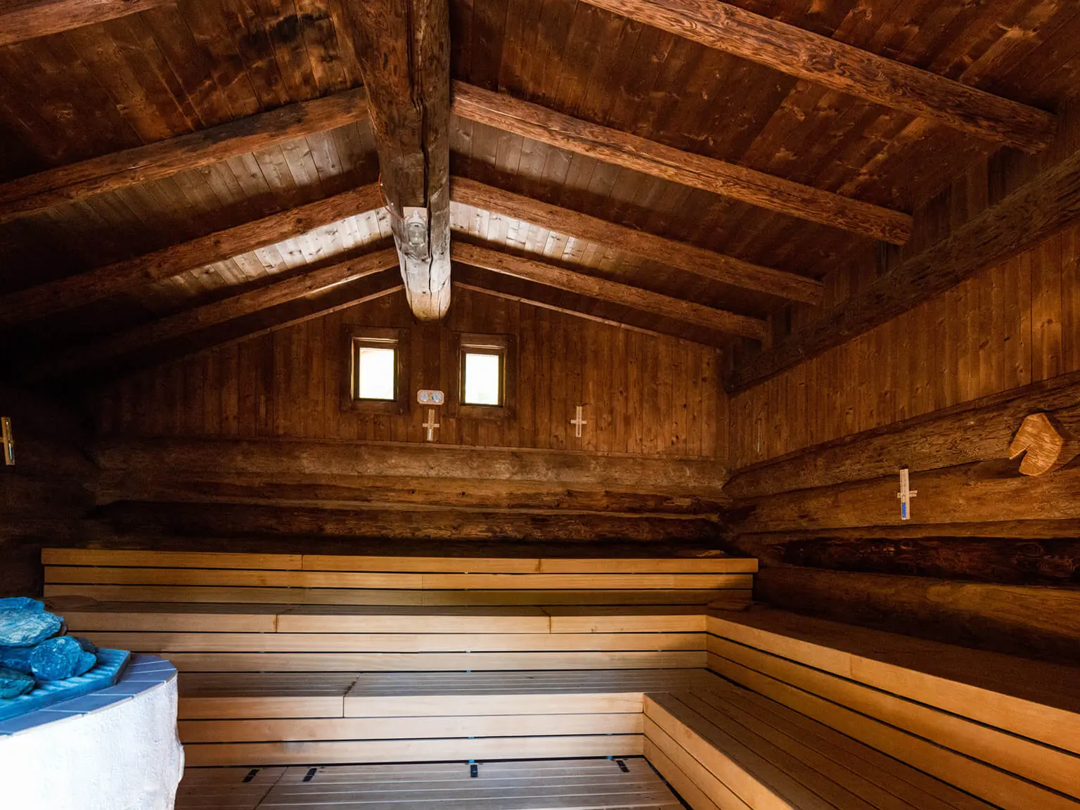 Innenansicht einer Sauna. Das helle Holz im mittleren und unteren Bereich sind Sitzbänke, das dunkle das Gebäude an sich. Vorne links ist ein Teil eines Aufgussofens zu sehen.